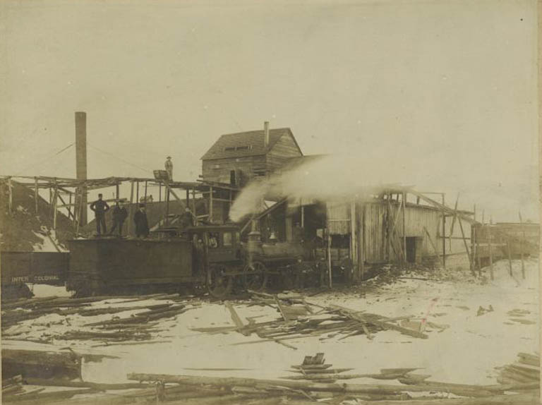 Photographie noir et blanc d’un bâtiment en bois avec trois hommes et une locomotive menée
