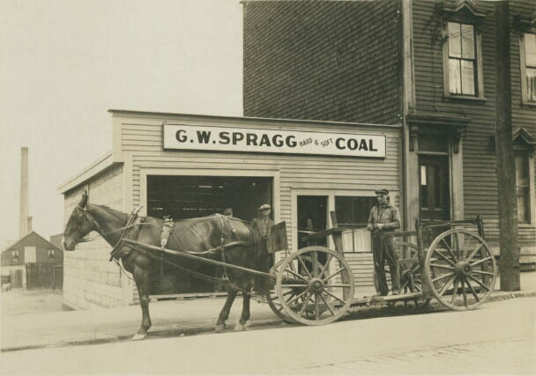 Photographie noir et blanc d’un cheval et d’un chariot avec trois hommes devant un bâtiment en bois