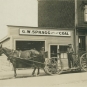 Photographie noir et blanc d’un cheval et d’un chariot avec trois hommes devant un bâtiment en bois