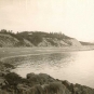 Photographie noir et blanc d’un rivage avec de hautes falaises et, au sommet, une maison