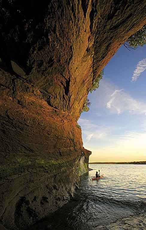 Colour photograph of a red rock cliff on the ocean with person in a kayak