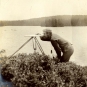 Photographie noir et blanc d’un homme devant un lac avec de l’équipement d’arpentage